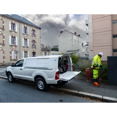 Véhicule utilitaire blanc garé, technicien en tenue de sécurité travaillant près d'une maison.